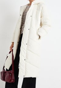 Padded knee-length coat in off-white with a hood, featuring a quilted design and snap buttons, paired with a burgundy shoulder bag.