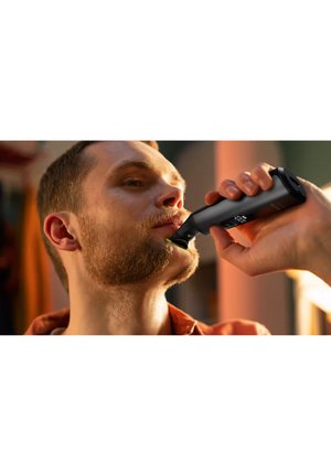 Mann schneidet Bart mit schwarzem elektrischen Trimmer drinnen, konzentriert sich darauf, den Bart in der Nähe des Kinns zu pflegen.