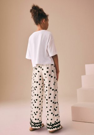 Young person with curly hair in a bun wears a white short-sleeve top and loose white pants with black polka dots and wavy trim.