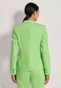 Grüner Blazer mit taillierter Passform, scharfen Revers und drei Knopfakzenten an den Ärmeln. Strukturierter Stoff sorgt für visuelles Interesse.