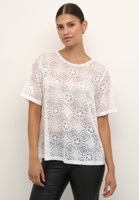 Weißes Spitzen-T-Shirt mit Blumenmustern, kurzen Ärmeln und einem runden Ausschnitt, über einem Oberteil in einfarbigem Design getragen. Glatte Textur und lockere Passform.