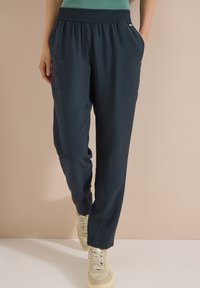 Person wearing dark blue elastic waist pants with front pockets and light-colored sneakers, standing against a beige background.