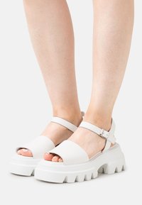 White open-toe sandals with a wide strap, adjustable ankle strap, and chunky textured sole. Smooth material with a matte finish.