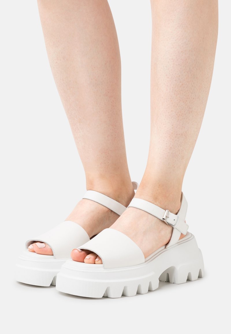 White open-toe sandals with a wide strap, adjustable ankle strap, and chunky textured sole. Smooth material with a matte finish.