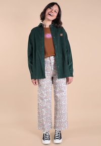 Chemise en velours côtelé vert à boutons, t-shirt graphique marron, pantalon large à motifs floraux, baskets noires à semelles blanches, texture lisse, coupe décontractée.