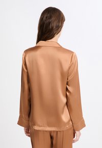 Satynowa, długa koszula do piżamy w ciepłym, jasnobrązowym kolorze. Posiada klasyczny kołnierzyk i gładką, błyszczącą fakturę. Rękawy wykończone mankietami.