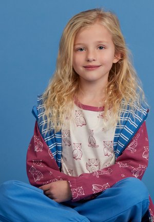 Kind dat een blauw gestreept hoodie draagt over een crème shirt met rode uilenprints en blauwe broek. Het haar is golvend en lichtblond.