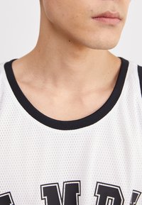Baltas sportinis marškinėlis su tinklelio tekstūra, su juodu rausvo kaklo apvadu ir ryškiais juodais užrašais priekyje. Detali nuotrauka.