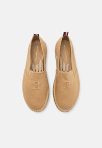 Des chaussures slip-on beiges avec une tige en toile, une semelle en jute tressé, un logo embossé et un onglet de talon rayé en marine, rouge et blanc.