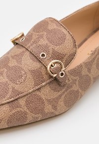 Gros plan d'un mocassin marron à motifs avec un tissu texturé et un détail de boucle métallique dorée sur le dessus.