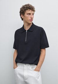 Mörk marinblå kortärmad polo med en platt krage och en frontdragkedjedetalj. Bärs med vita byxor, vilket framhäver en ren, minimalistisk design.