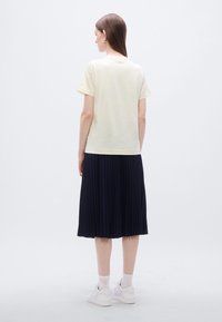 Ανοιχτό κίτρινο κοντομάνικο t-shirt συνδυασμένο με πλισέ ναυτικό μπλε φούστα. Το μοντέλο φοράει λευκά αθλητικά παπούτσια· απλός σχεδιασμός με λεία υφή.