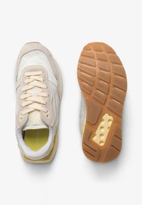 Beige en witte sneaker met crèmekleurige veters, weergegeven vanaf de bovenkant en zool, met een gestructureerde rubberen buitenzool in gumkleur en gele merkopdruk op de binnenzool.
