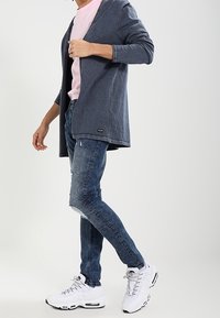 Dunkelblauer Cardigan, hellrosa T-Shirt, lässig zerstörte blaue Jeans und weiße Sportschuhe mit silbernen Akzenten. Legerer Stil mit Lagenlook.