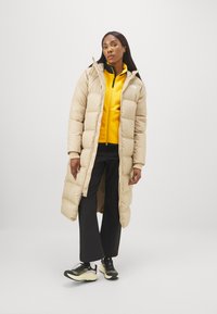 Longo cappotto imbottito beige con colletto alto e cerniera, indossato sopra una maglietta in pile gialla, pantaloni neri e scarpe da ginnastica nere.