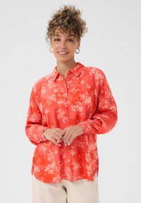 Frau mit einem Lächeln, lockigem, hochgestecktem Haar, die goldene Creolen trägt, ein korallfarbenes Blumenhemd mit Knöpfen und hellbeige Hosen vor einem weißen Hintergrund.