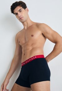 Schwarze Baumwoll-Boxershorts mit einem roten Bund, der das "TOMMY HILFIGER"-Logo trägt. Das Modell ist oberkörperfrei und posiert vor einem neutralen Hintergrund.