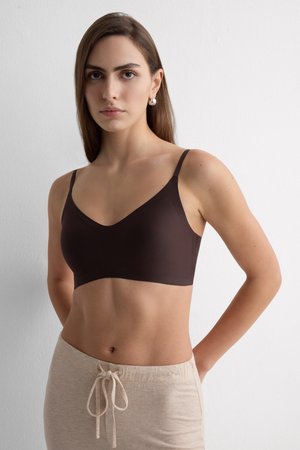 Frau mit glattem braunem Haar, die silberne Creolen, ein dunkelbraunes Crop-Top und beige Hose mit Kordelzug trägt und vor einer weißen Wand steht.
