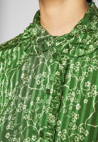 Camicia verde con colletto, caratterizzata da un motivo floreale con fiori bianchi, volant lungo il colletto e chiusura a bottoni frontale.