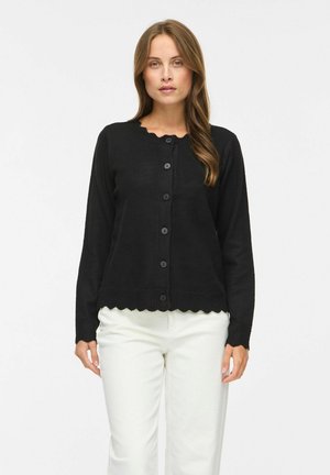 Femme portant un cardigan noir boutonné avec des bords festonnés et un pantalon blanc, debout devant un fond blanc uni.