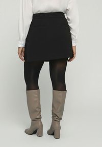 Schwarzer Minirock mit Gesäßtaschen, kombiniert mit schwarzen blickdichten Strumpfhosen und knielangen beige Lederschuhen mit einem robusten Absatz. Glatte Textur.