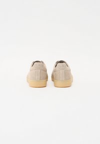 Lys grå semsket skinn sneakers med brune gummisåler. Har en rund form og sømbelagt detaljer. Merking synlig på hælen.