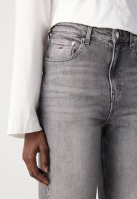 Jean gris taille haute sur une personne portant une chemise blanche à manches longues, la main gauche détendue le long du corps.