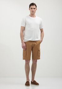 T-shirt blanc à manches courtes, en tissu léger ; shorts cargo marron avec deux poches latérales ; chaussures brunes à enfiler. Tenue simple et fonctionnelle.