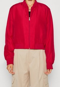 Bijgesneden rode bomberjack van glanzende stof, met ritssluiting en elastische manchetten. Gecombineerd met losse beige broek.