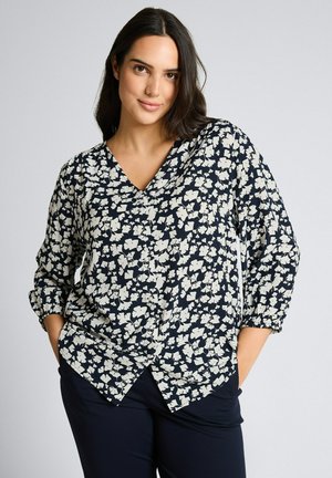 Frau mit dunklen Haaren, die eine schwarz-weiße, langärmelige Bluse mit floralem Muster und schwarze Hosen trägt, steht mit den Händen in den Taschen vor einem einfarbigen Hintergrund.