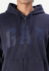 Persona che indossa una felpa con zip di colore blu navy, con un grande logo "GAP" sul petto e lacci visibili sul cappuccio.