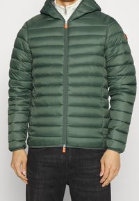 Grøn pufferjakke med hætte, der har vandret quilting, lynlås lukning og orange lynlåstræk. Lavet af skinnende, glat materiale.