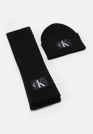 Écharpe en tricot côtelé noire et bonnet avec étiquettes du logo Calvin Klein Jeans sur fond blanc.