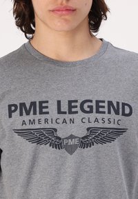 Šedé bavlněné tričko s tmavým tištěným logem s křídly a textem "PME LEGEND AMERICAN CLASSIC." S kulatým výstřihem a krátkými rukávy.
