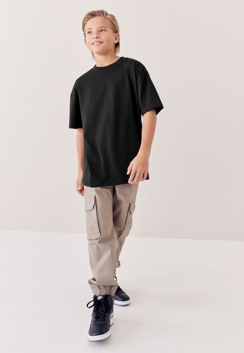 Niño con camiseta negra de gran tamaño, pantalones cargo beige y zapatillas negras caminando con una ligera sonrisa sobre un fondo liso.