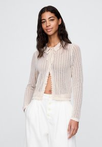 Langärmelige, offene Strickjacke in hellem Beige, mit einem Strickmuster, Rüschensaum und einer Schleife am Kragen.