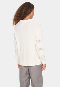White knitted sweater with long sleeves featuring lace stripes. The back has a relaxed fit and ribbed hem, paired with gray trousers.