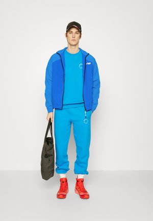 Zila sporta jaka virs zila krekla un biksēm, apvienota ar sarkaniem kedām. Melna cepure un pelēka soma ar rūtainu rakstu.