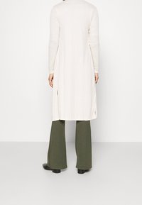 Cárdigan largo de canalé en un tono beige claro, con mangas largas, aberturas laterales y un corte relajado, combinado con pantalones de campana en color verde oliva y botines negros.