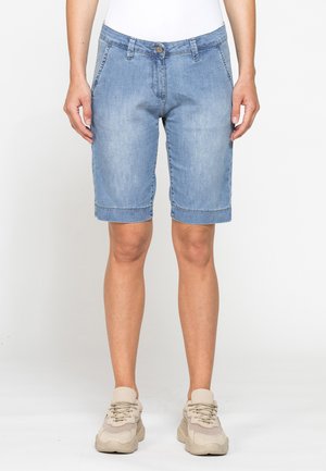 Persona che indossa pantaloncini di jeans azzurri fino al ginocchio, top bianco e sneakers beige, in piedi davanti a uno sfondo bianco uniforme.