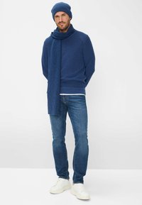 Herenoutfit met een donkerm blauwe gebreide trui, bijpassende beanie, geribde sjaal en lichtblauwe spijkerbroek met witte sneakers.