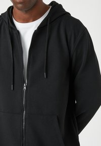 Sudadera negra con cremallera y fabricada en un tejido texturizado, con bolsillos frontales y cordones en la capucha. Presenta un diseño acanalado y una cremallera plateada.