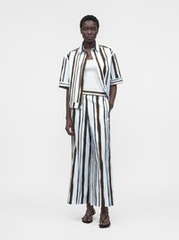 Femme portant une chemise ample rayée assortie et un pantalon à jambes larges avec des sandales noires et un débardeur blanc sur fond uni.
