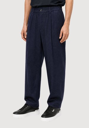 Uomo che indossa pantaloni larghi blu navy con vita elastica e scarpe da ginnastica nere con suola bianca, in piedi su uno sfondo semplice.