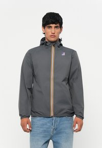 Chaqueta cortavientos gris con capucha, que presenta un cierre frontal con rayas naranja y blancas, y un logo en el lado izquierdo del pecho.