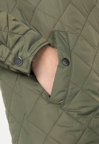 Mano parcialmente dentro del bolsillo delantero de una chaqueta acolchada verde con detalles de botones a presión en el puño y en la solapa del bolsillo.