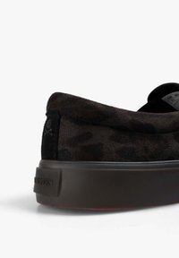 Zapatilla baja de ante negra con un sutil patrón de camuflaje, suela de goma elevada y un pequeño logo de calavera y huesos cruzados en la lengüeta del talón.