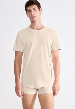 Jonge man draagt een beige korte mouwen t-shirt en bijpassende boxershorts, staand tegen een effen witte achtergrond.