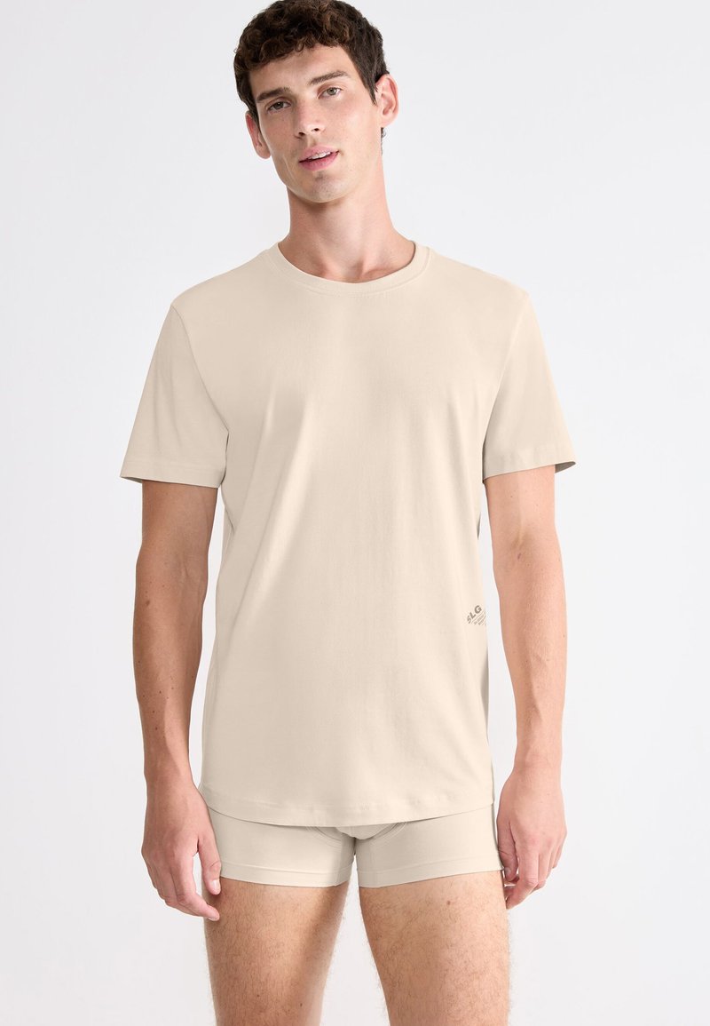 Jeune homme portant un t-shirt beige à manches courtes et des boxers assortis, debout devant un fond blanc uni.