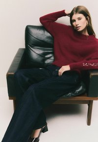 Burgundy stickad tröja med texturerad yta och dekorerade ärmavslut, kombinerat med mörka denimbyxor med vida ben; sitter på en svart läderstol.